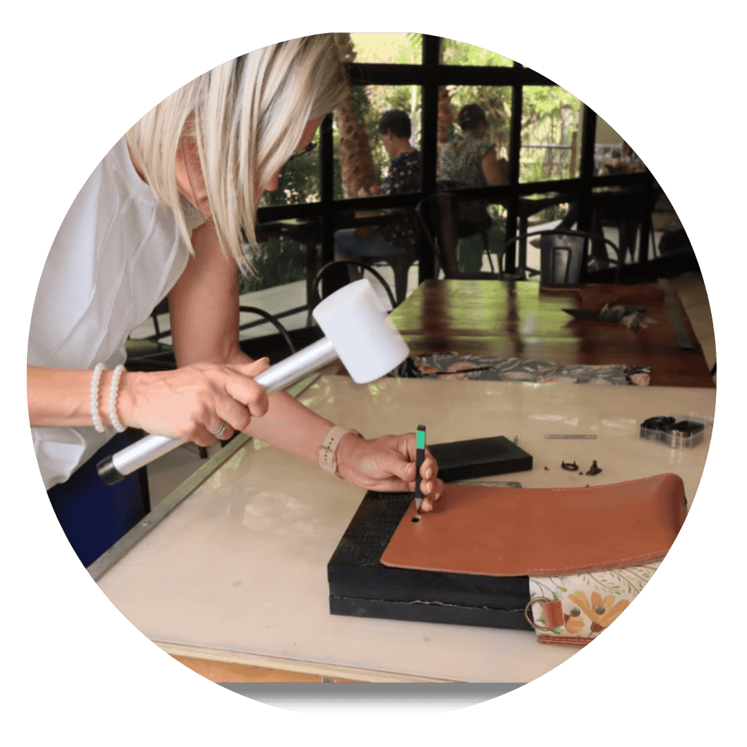 A woman working on leather craft using a hammer and punch tool to shape a brown leather piece during a hands-on workshop.